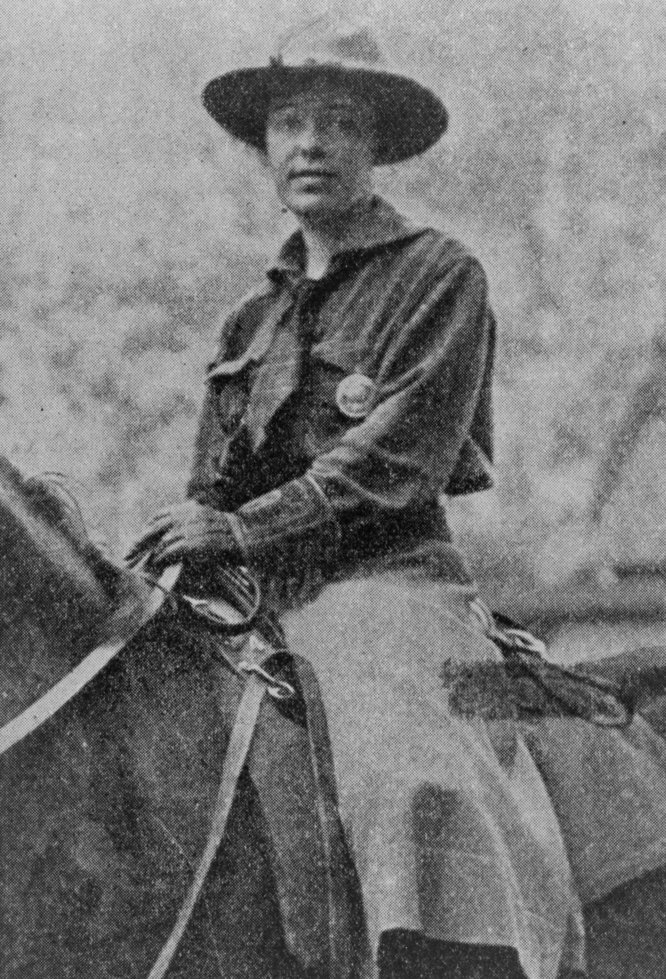 Clare Hodges on horseback wearing a split skirt, shirt, bandana, gloves, and a flat brim hat with a round badge pinned on the pocket.
